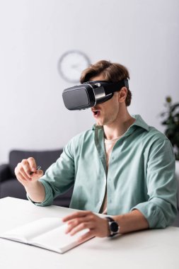 Şok olmuş bir adamın seçici odak noktası VR kulaklıklı elinde kalem, masanın üzerinde defter var. 