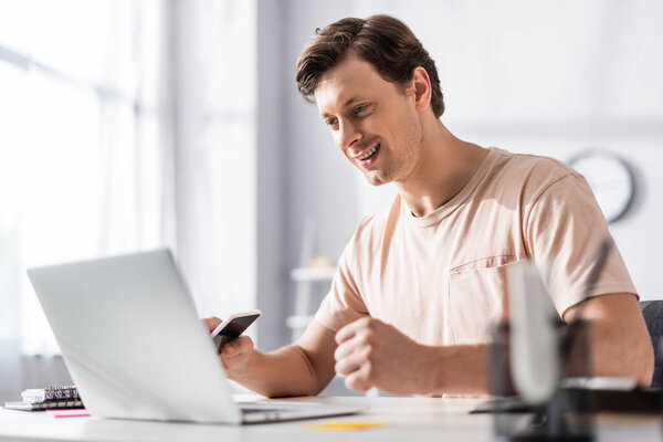 Selective focus of smiling freelancer looking at laptop screen and holding cellphone at home, earning online concept