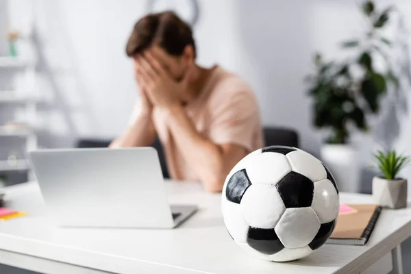 Selective focus of football on table and sad man covering face with hands near laptop at home, earning online concept