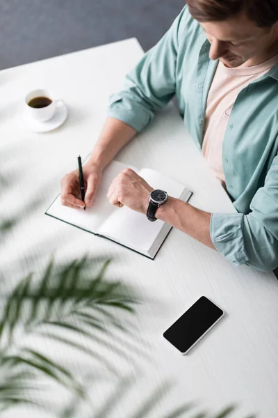 Foco Seletivo Freelancer Verificando Tempo Enquanto Escreve Notebook Perto Smartphone — Fotografia de Stock