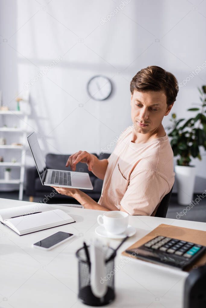 Selective focus of concentrated freelancer holding laptop and sitting near table with stationery at home, earning online concept