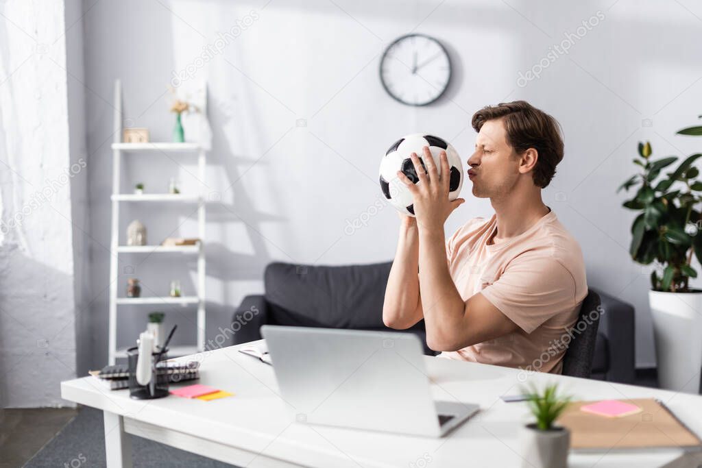 Selective focus of man kissing football while sitting near laptop and stationery on table at home, concept of earning online