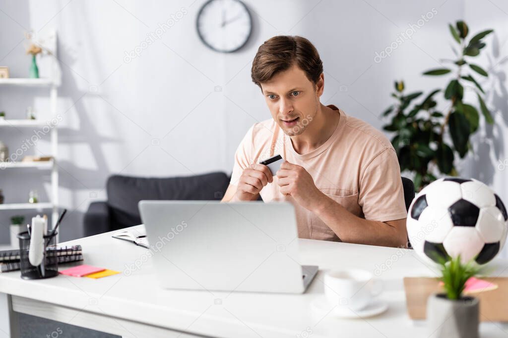 Selective focus of man holding credit card and looking at laptop near football and stationery on table, concept of earning online