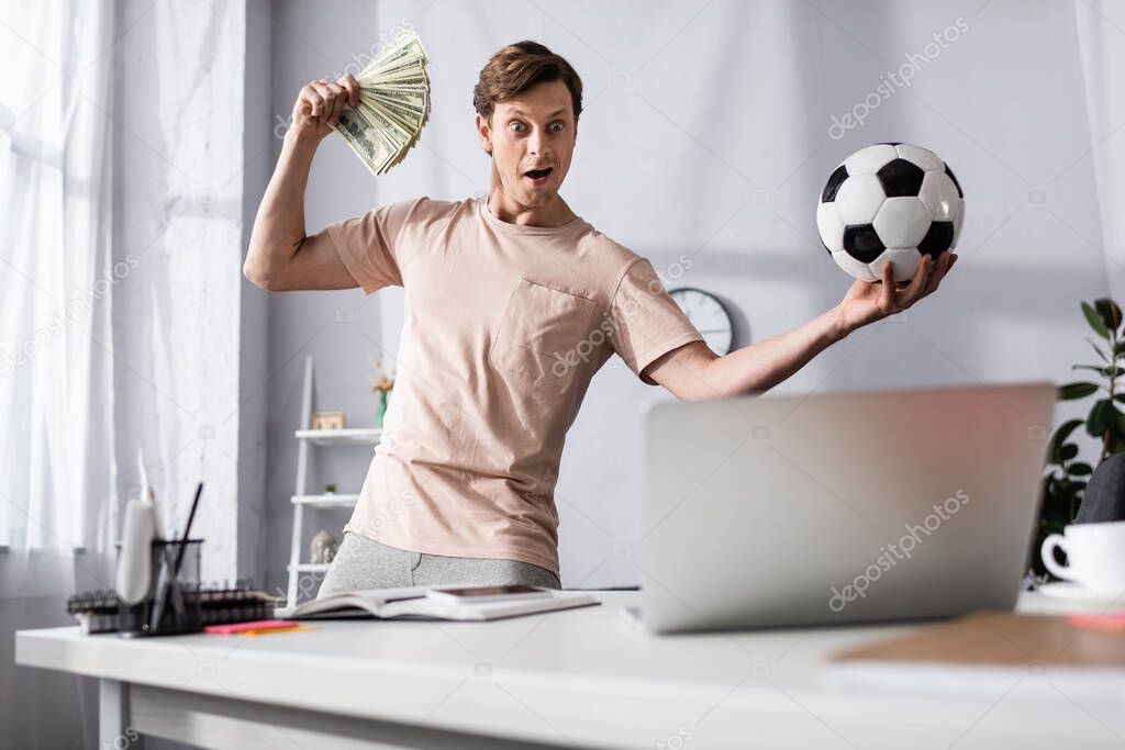 Selective focus of shocked man with cash and football looking at laptop in living room, concept of earning online