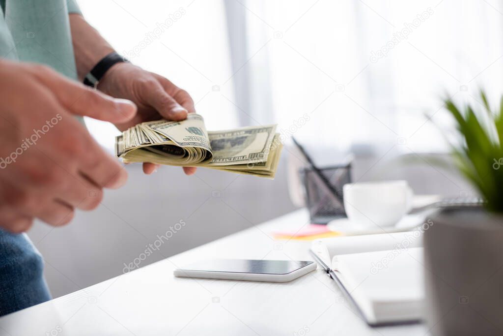 Cropped view of young man counting dollar banknotes near smartphone with blank screen and notebook on table, earning online concept