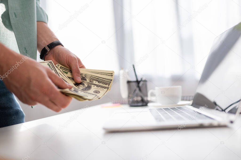 Selective focus of young man counting dollars near laptop and cup of coffee on table, earning online concept