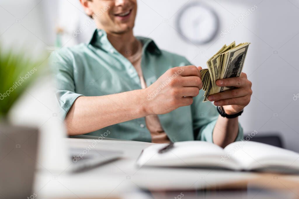 Cropped view of smiling freelancer counting dollars near laptop and notebook on table, earning online concept