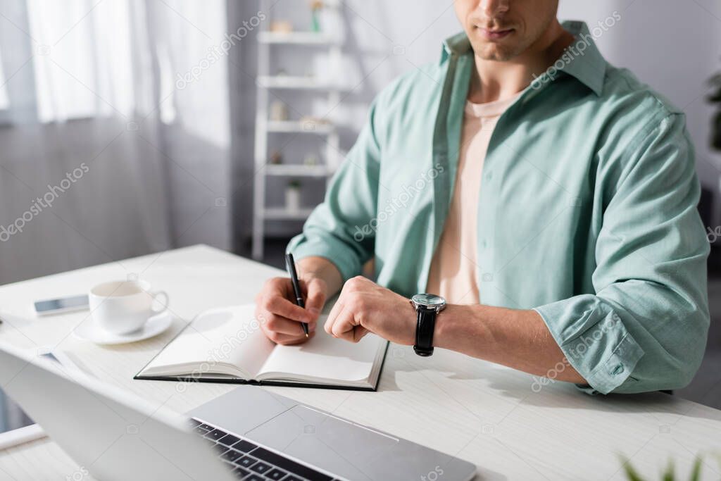 Cropped view of freelancer checking time on wristwatch while working with notebook and laptop at home, concept of time management 