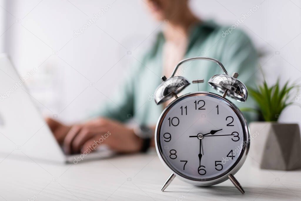 Selective focus of alarm clock on table near man using laptop at home, concept of time management 