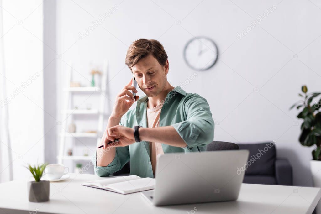 Selective focus of man checking time while talking on smartphone near laptop and notebook on table, concept of time management 