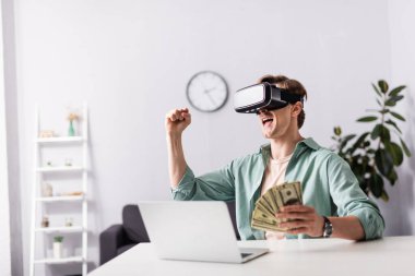 VR kulaklıklı neşeli adam, evet jest yapıyor ve parayı masanın yanında tutuyor, online konsept kazanıyor.