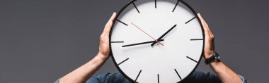 Panoramic crop of man holding clock near face isolated on grey, concept of time management  clipart