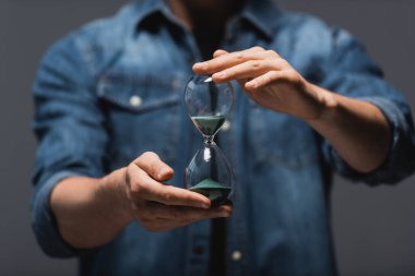 Cropped view of man holding hourglass with flowing sand isolated on grey, concept of time management  clipart