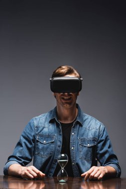 VR kulaklıklı gülümseyen adamın seçici odağı gri arka planda, kum saatinin yanında oturuyor, zaman yönetimi kavramı. 