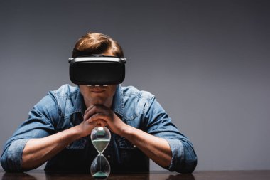VR kulaklıklı adam gri arka planda, kum saatinin yanında oturuyor, zaman yönetimi kavramı. 