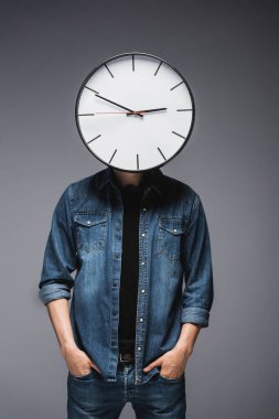 Young man with clock on head and hands in pockets of jeans on grey background, concept of time management  clipart
