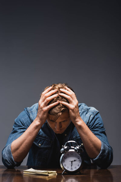 Депрессивный человек сидит рядом с деньгами и будильником на столе на сером фоне, концепция управления временем 