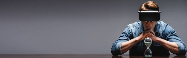 Panoramic crop of young man in vr headset sitting near hourglass on table on grey background, concept of time management 