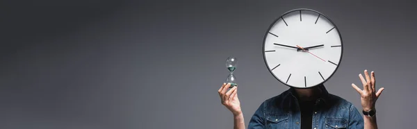 頭の上に時計を持つ男のパノラマショット砂時計を保持し 灰色の背景にジェスチャー 時間管理の概念 — ストック写真