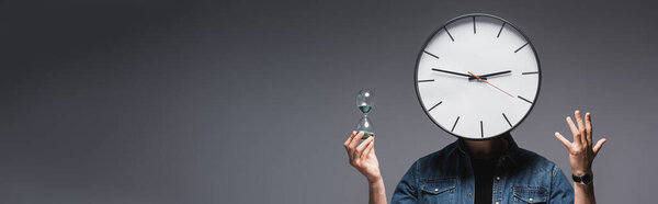 Panoramic shot of man with clock on head holding hourglass and gesturing on grey background, concept of time management 