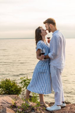 Mutlu kadın ve adamın kırmızı şarapla bardakları tuttuğu ve gölün yanında durduğu yan manzara.