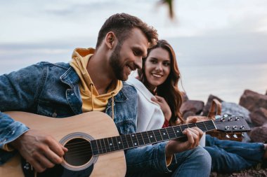 Sakallı adamın akustik gitar çaldığı seçici odak noktası kız arkadaş ve şenlik ateşi.