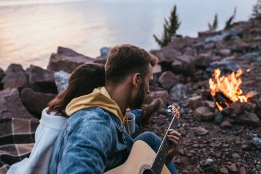 Akustik gitar çalan sakallı adamın seçici odak noktası kız arkadaşının yanında ve şenlik ateşine bakıyor.