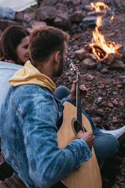 Sakallı adamın akustik gitar çaldığı seçici odak noktası kız arkadaş ve şenlik ateşi.