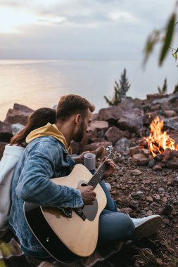 Akustik gitar çalan sakallı adamın seçici odak noktası kız arkadaş, nehir ve şenlik ateşi.