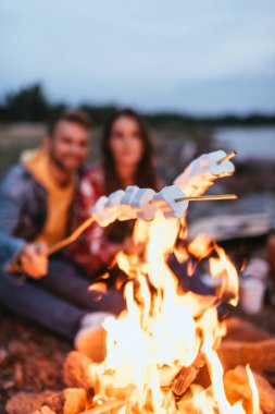 selective focus of puffy marshmallows on sticks near burning bonfire and couple clipart