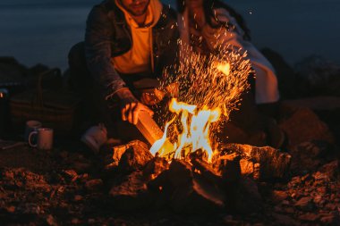 cropped view of man putting log in bonfire near girl  clipart