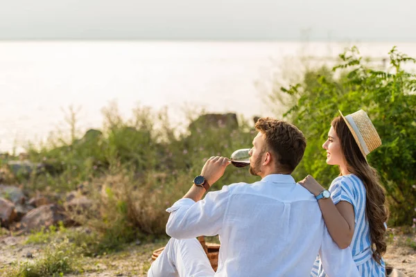 湖の近くで赤ワインを飲む男に触れる麦藁帽子の陽気な女性 — ストック写真