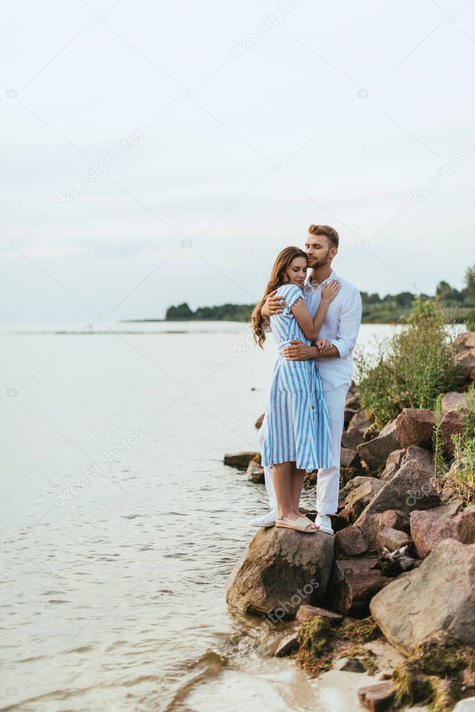 handsome man hugging beautiful woman in dress standing on rocks near river 