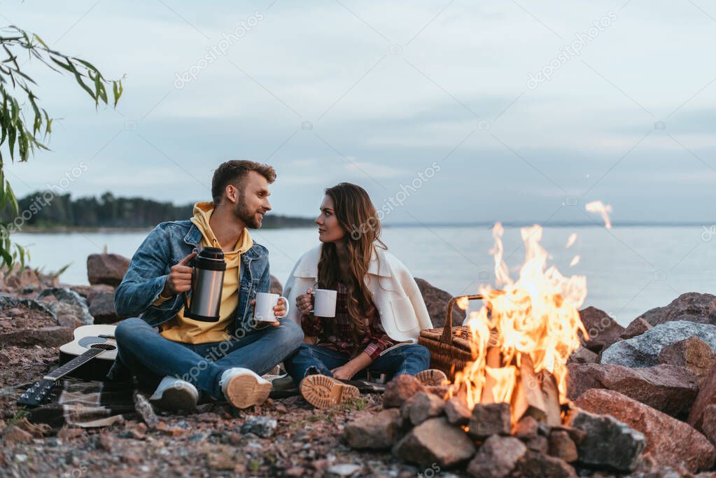 selective focus of couple holding cups and sitting on rocks near bonfire and lake
