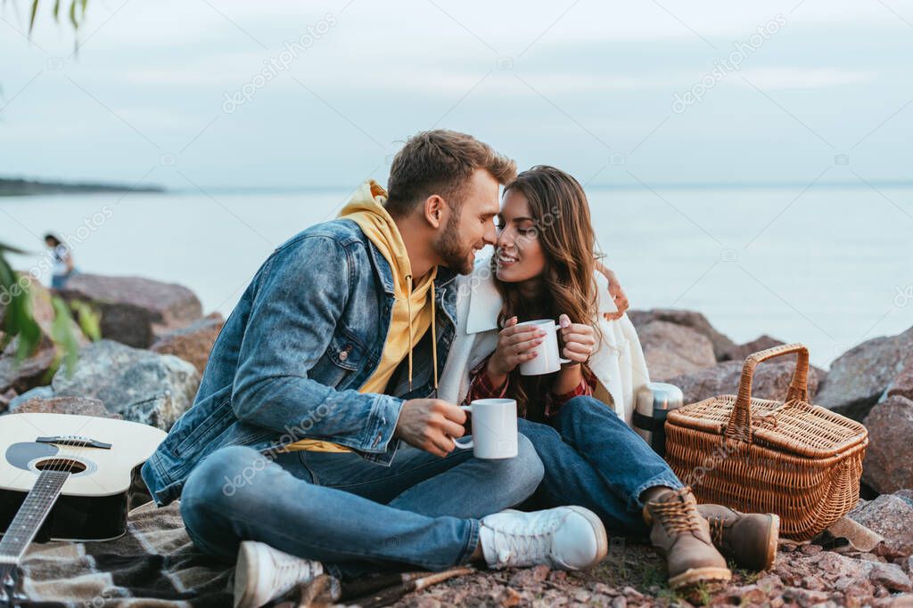happy couple holding cups and sitting on blanket near river