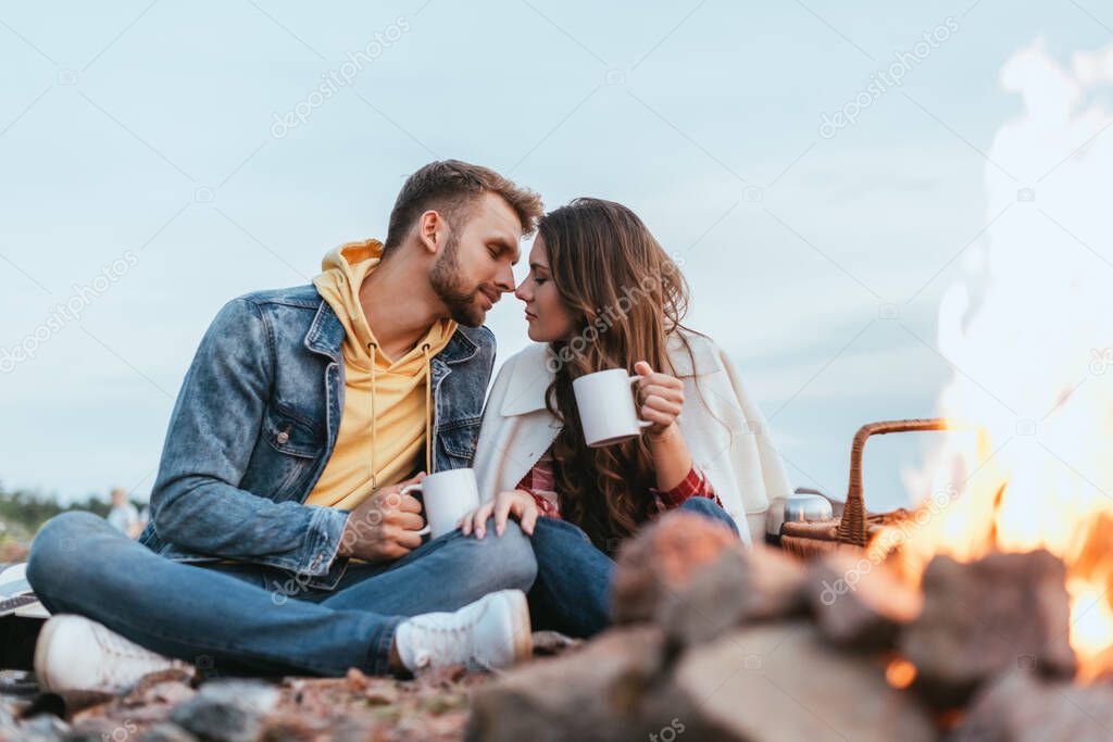 selective focus of handsome man and attractive woman holding cups of tea near bonfire