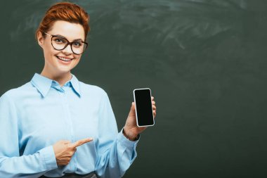 Gözlüklü mutlu öğretmen karatahtanın yanındaki boş ekranla akıllı telefonu işaret ediyor. 
