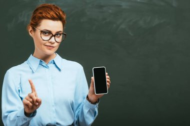 Şüpheci öğretmen, akıllı telefonu karatahtanın yanında boş ekranla tutarken yasaklayıcı bir hareket gösteriyor