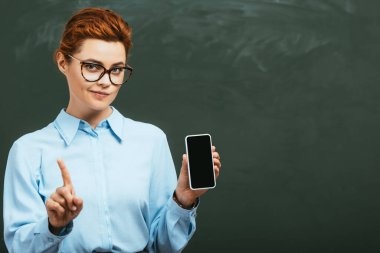 skeptical teacher showing forbidding gesture while holding smartphone with blank screen near chalkboard