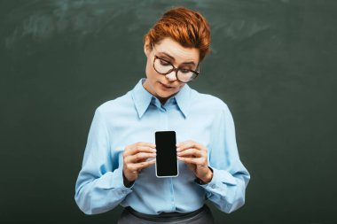 Güzel genç öğretmen kara tahtanın yanında duruyor ve elinde boş ekranla akıllı telefonu tutuyor.
