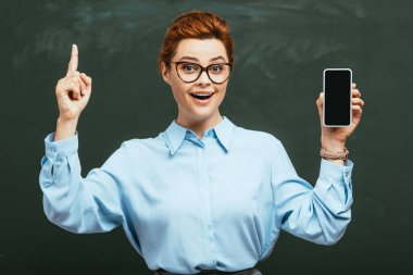 Gözlüklü neşeli öğretmen elinde kara tahtanın yanında boş ekranlı akıllı telefon tutuyor.   