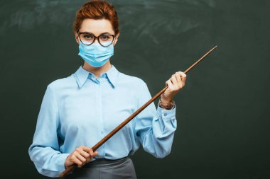Tıp maskeli genç öğretmen boş karatahtanın yanındaki sopayı tutarken kameraya bakıyor. 