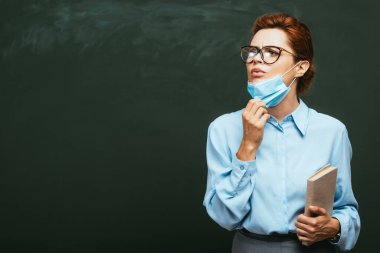 Tıp maskesine dokunarak tahtaya yakın duran ve gözlerini kaçıran bir öğretmen. 