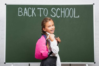 cheerful schoolgirl pointing with thumb at backpack near chalkboard with back to school lettering clipart