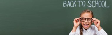 horizontal image of excited schoolgirl laughing with closed eyes and touching eyeglasses near chalkboard with back to school text clipart