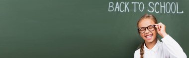 panoramic crop of excited schoolgirl laughing with closed eyes and touching eyeglasses near chalkboard with back to school lettering clipart