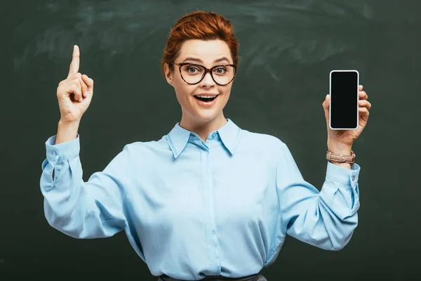 戴眼镜的快乐老师 在黑板旁边拿着装有空白屏幕的智能手机 — 图库照片