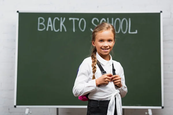 有选择性地聚焦在可爱的女生身上 背负盈亏地微笑在黑板旁边的相机前 背对着学校的题词 — 图库照片