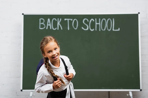 学校の碑文に戻ると黒板の近くのカメラで笑顔バックパックを持つ陽気な女子高生 — ストック写真