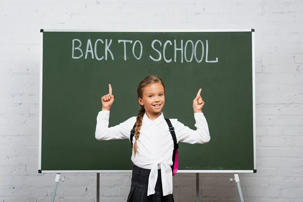 Fröhliche Schülerin Zeigt Mit Den Fingern Auf Schulschriftzug Auf Kreidetafel — Stockfoto